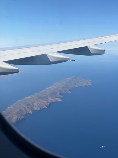 A view from an airplane window shows an island in the ocean. The wing of the plane is visible on the left side of the frame, and the island, which is rugged and mountainous, is situated in the center. The ocean surrounds the island, and the sky is clear and blue. A small boat is visible in the distance. The location is within the vicinity of the Paiania Municipal Unit, Greece.