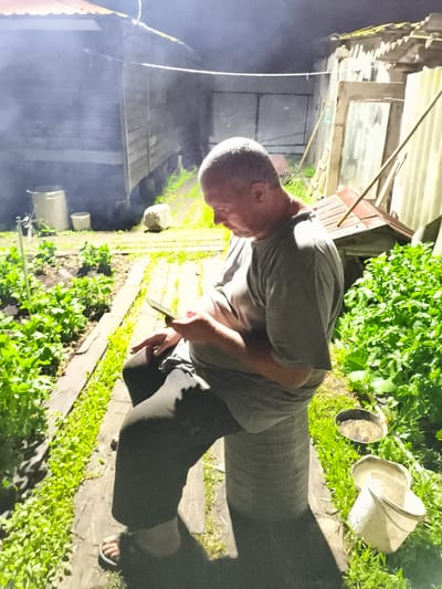 A middle-aged man with short, graying hair is sitting outdoors at dusk in what appears to be a rural setting in Il'mino, Russia. He is wearing a gray t-shirt and dark pants, and is looking down at a smartphone in his hands. The man is seated on a cylindrical stool. In the background, there is a wooden building with a corrugated metal roof and a fence. There is also dense green vegetation, possibly a garden, and some buckets. Smoke is visible in the air, suggesting that a fire or cooking may be taking place nearby. The lighting suggests it is either early morning or late evening.