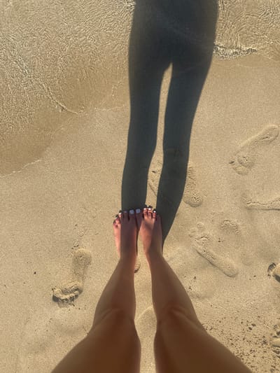 A first-person perspective shot of a person's bare feet and lower legs standing on a sandy beach. The feet are adorned with white-painted toenails and are positioned in the foreground. Behind the feet, footprints are visible in the sand, some partially submerged in shallow, clear water with a rippled surface reflecting sunlight. A long shadow of the person's legs is cast across the sand and into the water. The scene is located on a beach in Protaras, Cyprus, identifiable by the characteristic golden sand and the crystal-clear waters of the Mediterranean Sea.