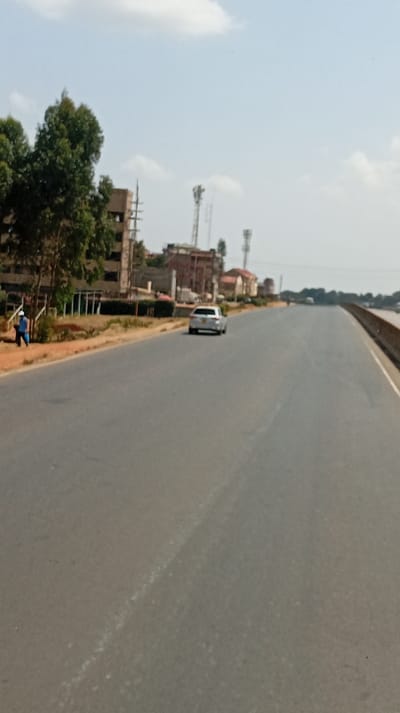 The image depicts a multi-lane asphalt highway in Sigona ward, Kenya, under a clear sky with scattered white clouds. The road, which curves slightly to the right in the distance, has a faint white dashed line indicating lanes. A single white sedan is visible traveling away from the camera in the rightmost lane. On the left side of the road, an unpaved reddish-brown shoulder runs alongside a row of green trees and various buildings. One individual, wearing a blue top and light-colored bottoms, with a possible light-colored head covering, is walking away from the camera on this shoulder, carrying a blue object in their left hand. In the background on the left, several multi-story buildings, some appearing to be under construction, are visible behind the trees. Multiple tall metallic communication towers are also prominent in the mid-background. A guardrail or barrier runs along the right side of the road in the distance, suggesting an elevated section. The scene is illuminated by daylight.
