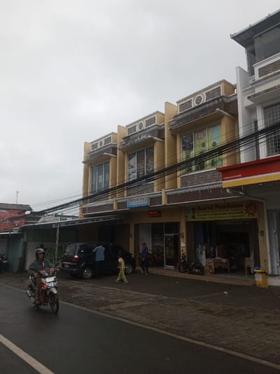 A street scene reveals a row of two-story buildings, the center ones painted a tan color with decorative stone accents. A black van is parked in front of one of these buildings where a man seems to be working on the car. A child walks near the van. An individual on a moped rides by on the left side of the frame. Above, a dense cluster of electrical wires cuts across the sky. The weather appears overcast.