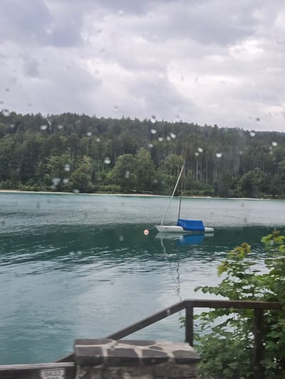 The image depicts a serene lake scene, likely Kochel am See, Germany, under an overcast sky. The water is a deep teal, with subtle ripples indicating calm conditions. In the middle ground, a small white sailboat is moored, its blue cover draped over the boom. A single orange buoy floats nearby. The background is dominated by a dense, dark green forest that stretches along the shoreline and up the rolling hills. In the foreground, the edge of a wooden railing and stone structure are visible, suggesting the vantage point is from a dock or elevated pathway. Numerous water droplets are scattered across the lens, creating a bokeh effect that slightly obscures the details of the scene. The overall atmosphere is tranquil and slightly melancholic due to the cloudy weather.
