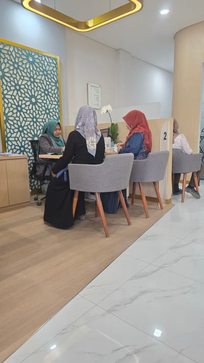 In the city of Blangpeuria, Indonesia, a waiting area is depicted with several individuals present. A woman wearing a green hijab and a grey jacket is seated at a desk, appearing to be engaged in a conversation or transaction with another person. To her right, a woman in a grey hijab and a long black dress sits in a grey armchair, facing away from the camera. Further to the right, another woman in a red hijab and a blue top is seated in a similar grey armchair, also facing away. In the background, a fourth woman, partially visible, is seated in a grey armchair with her back to the camera. The environment features a decorative wall panel with an intricate geometric pattern, a white orchid plant, and a floor that transitions from wood-look laminate to white marble tiles. A desk with a laptop is visible, along with a sign that reads "FLOOR DIRECTORY." The overall scene suggests a service or administrative setting.
