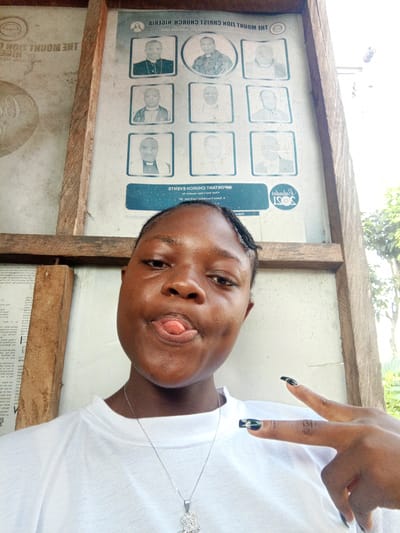 A young person with dark skin and braided hair is featured prominently in a close-up selfie, making a playful expression with their tongue slightly out. They are wearing a plain white t-shirt and a silver necklace with a decorative pendant. Their left hand is raised, forming a 'V' or peace sign, with long, dark, and possibly patterned fingernails visible.
The background consists of a wooden-framed structure supporting a large display board. The board is light-colored with various elements printed on it. On the right side, there are several small framed photographs of men, likely clergy, arranged in a grid pattern. Much of the text on the board appears upside down and reversed due to the photo being taken with the phone inverted or the board itself being seen through a reflection or from an unusual angle. Legible text includes phrases like "THE MOUNT ZION CHRIST CHURCH NIGERIA" and "IMPORTANT CHURCH EVENTS," indicating a religious context. A logo or seal with "2021" is also visible. The left side of the board contains more printed text, which is largely faded or obscured but seems to be informational notices or scriptures.
The overall setting appears to be outdoors or in a semi-open area, suggested by the bright natural light and some foliage visible in the upper right background. The general atmosphere is casual and lighthearted. The location is Mbribit, Nigeria, as provided.