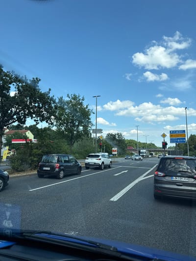 This is a vertical, slightly angled, eye-level shot taken from inside a vehicle, likely a car, looking outwards at a highway intersection. The scene depicts a traffic jam on a sunny day. The sky is a vibrant blue, dotted with fluffy white clouds, suggesting good weather. The road is asphalt with clear white lane markings, and several cars are stopped in traffic.

Prominently displayed are German road signs, indicating directions to various cities. One sign shows routes to Kassel, Marburg, Büdingen, Frankfurt, Dortmund, and Wetzlar, with route numbers like 485 and 457. Another sign displays the route number 485 with an arrow pointing right. The license plates of two cars are visible: "LDK BM 318" and "MR FA 2019". The presence of German license plates and road signs strongly suggests the location is Germany, consistent with the Giessen context.

On the left side of the road, there are trees and greenery, with some buildings visible in the distance, including a house with a red-tiled roof and a building with red and white sections. The overall impression is a typical highway scene during a traffic slowdown on a clear, bright day. The angle of the shot and the presence of other vehicles create a sense of being part of the traffic flow, looking out at the road ahead.