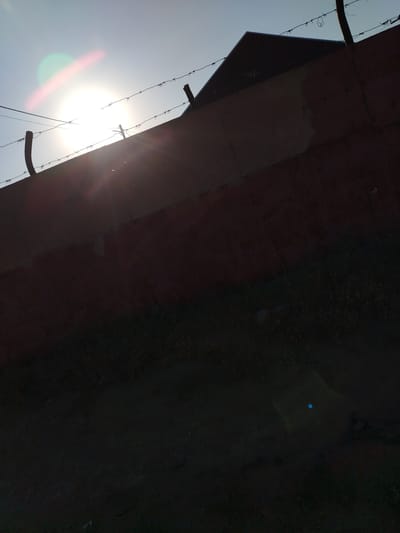The image shows a bright sun over a rusted metal fence topped with barbed wire in Töretam, Kazakhstan. A pointed structure, possibly a roof, is visible behind the fence. The foreground is dark and appears to be ground with some sparse vegetation. Lens flare from the sun is prominent in the upper left and lower right of the image.