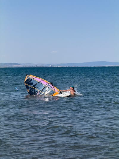 A man wearing an orange cap is windsurfing in the sea near Atiya, Bulgaria. The windsurfing sail is submerged in the water, and the man is attempting to right it. A plastic bottle is floating in the water to the left of the sail. The water is blue and wavy, with a distant coastline visible on the horizon. The sky is clear and blue.