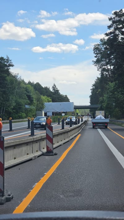 The image shows a view from a vehicle on a German highway, likely near Calau, Germany, given the context. The scene depicts a sunny day with scattered white clouds in a bright blue sky. The highway has multiple lanes, marked with white and orange lines. Construction barriers and orange warning lights are visible along the side of the road, indicating road work is in progress.
Several vehicles are present on the highway, including a white truck with a load covered in white plastic, and several passenger cars, including a dark colored Audi. The surrounding area is heavily wooded with tall green trees. A bridge or overpass is visible in the distance. The time of day appears to be daytime, possibly mid-morning or afternoon, due to the bright sunlight and clear skies. No people are clearly visible, but the presence of vehicles suggests traffic. The image captures a typical highway scene with a focus on the road and surrounding environment during what seems to be a period of construction.