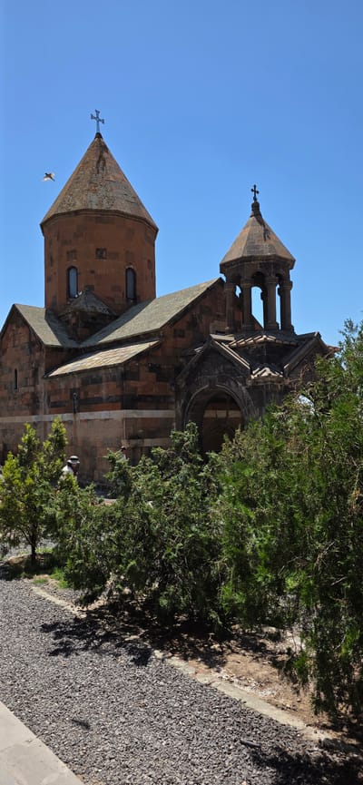 In Lusarat, Armenia, on a clear, bright day with a vibrant blue sky, an ancient church stands with its characteristic conical roof and bell tower. The church is constructed of reddish-brown stone, accented with darker bands, and features arched windows and a prominent cross atop its main dome. To the right, a smaller bell tower with an octagonal roof and open arches is visible. In the foreground, lush green trees and shrubs partially obscure the view of the stone pathway leading to the church. Two individuals, a person wearing a hat and another partially visible figure, can be seen near the church entrance, engaged in what appears to be a leisurely stroll. A bird is captured mid-flight in the upper left quadrant of the image, adding a dynamic element to the serene scene. The ground in the foreground is covered with dark gray gravel.