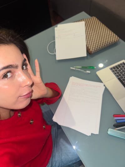 An overhead, high-angle shot captures a person partially visible, looking up, with a table spread with work-related items in the foreground. The person's face, from just above the forehead to the chin, and part of their upper body are visible. They have dark hair, prominent eyebrows, and wide, brown eyes looking upwards, with their skin appearing luminous, catching the light. They are wearing a red long-sleeved top with three visible round, light-brown buttons, and part of their denim-clad legs can be seen. Their hand is near their face, with fingers spread slightly.
The table, which appears to be made of light grey or blue glass, is cluttered with various objects suggesting work or study. These include a silver laptop with its keyboard visible, a stack of white papers (some with blurry red and black text), two pens (one green, one white/clear), and a small, patterned brown and beige woven bag or case. A container holding more pens or pencils and a long, clear object are also on the table.
The arrangement suggests a paused work or study session. The person's upward gaze and relaxed posture imply a moment of reflection or a break from their activity. The reflections on the glass table and laptop suggest an indoor setting, likely under artificial lighting. No specific outdoor weather or time of day is discernible. Text is present on the papers but is too indistinct to be read.