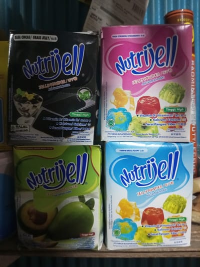 The image displays four boxes of Nutrijell jelly powder arranged in a stack, two on top and two on the bottom, on a wooden surface. The boxes are of different flavors: the top left box is black and labeled "Nutrijell Grass Jelly", the top right box is pink and labeled "Nutrijell Strawberry", the bottom left box is green and labeled "Nutrijell Avocado", and the bottom right box is blue and labeled "Nutrijell Plain". The background consists of a blurry blue and grey surface, possibly a wall or shelving. There are no people or animals visible in the image. The setting does not provide any specific clues to pinpoint a location within the city of Turumame, Indonesia, other than the text on the boxes indicating production in Indonesia.