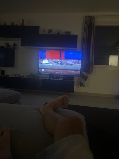 The image captures a living room interior in Arre, Italy, with a focus on a television screen displaying "The Amazing Digital Circus." The scene is dimly lit, suggesting it is evening or nighttime. A person's legs and feet are visible in the foreground, positioned on a sofa. The television, mounted on a dark entertainment unit, shows a colorful cartoon with characters. Various decorative items and electronics are placed on shelves and the entertainment unit. A window with a curtain is visible in the background. The floor appears to be made of wood planks.