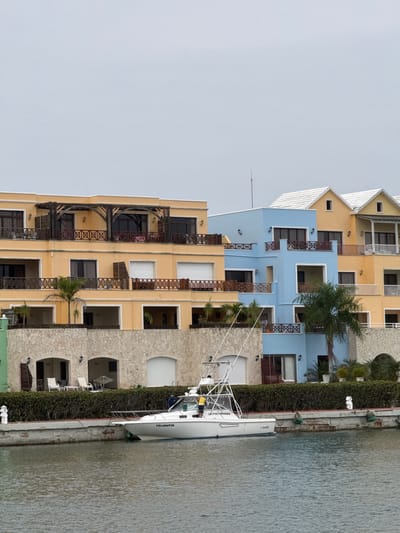 The image shows a waterfront scene in the city of Higüey, Dominican Republic. In the foreground, a white fishing boat with the name "YELLOWFIN" on its side is docked at a concrete pier. The boat has a tall antenna and fishing equipment on its deck, and two people are visible on board. Behind the pier, a dense green hedge lines the waterfront.
Further back, a series of multi-story buildings with colorful facades are visible. The buildings are predominantly yellow and light blue, with balconies and wooden railings. The architecture suggests a tropical or Mediterranean style, with arched doorways and stone accents on the lower levels. Palm trees and other tropical vegetation are interspersed among the buildings. The sky is overcast, suggesting a cloudy day. The overall scene depicts a resort-like or residential area along a waterway.