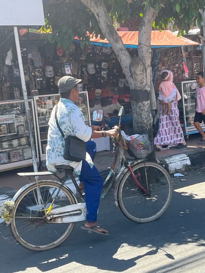 In the image, a man wearing a cap, a floral shirt, and blue pants rides a bicycle on a street in Desa Tambakreja, Indonesia. He has a dark crossbody bag on his shoulder. The bicycle has a basket in the front containing a white bag. To the right of the man, a woman in a pink patterned dress holds a child wearing pink. Further to the right, a young boy in a pink t-shirt walks. Behind them, shops display various items including hats and accessories. The scene is set under a tree with leafy branches, and the ground is paved. The lighting suggests it is daytime, with shadows visible on the road.