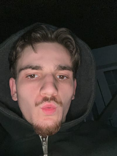 A young man with brown eyes and short, wavy brown hair poses in a dark grey hoodie. He has a light beard and mustache. His lips are slightly pursed. The background is mostly dark, with a possible structural element visible behind him. The image is taken from a slightly high angle, tilted to the side, focusing on the man's face.