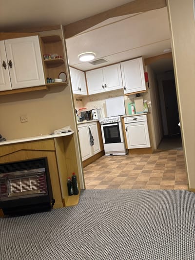 The image shows the interior of a small, possibly mobile, home. A gas fireplace is visible in the foreground, set into a wooden surround. To the right, a doorway leads into a kitchen area. The kitchen features white cabinets, a white oven with a black door, and a countertop with various items including bottles and a cutting board. The flooring in the kitchen is tiled in a checkerboard pattern of brown and beige squares. A patterned carpet covers the floor in the area with the fireplace and extends into a hallway or passage visible in the background. The overall impression is of a modest and functional living space. The location is identified as Wargrave, United Kingdom.