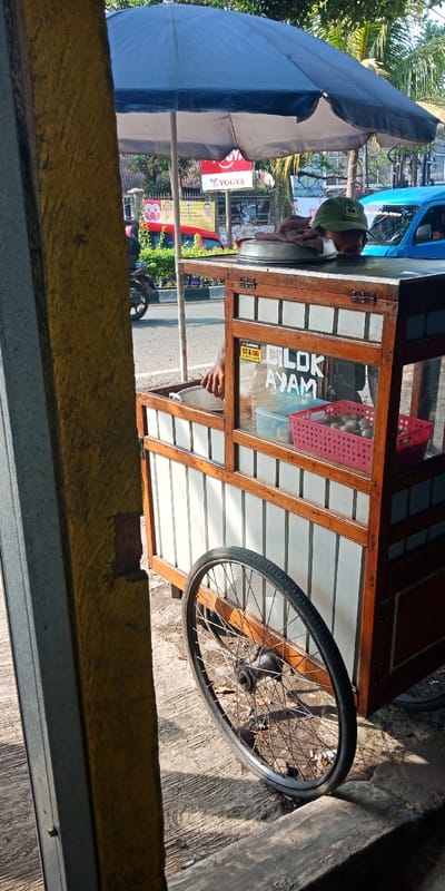 The scene depicts an outdoor street vendor operation in Bojongherang, Indonesia, under bright daylight. A vendor, partially visible from the chest down, wearing a green cap, is positioned behind a wooden and glass mobile food cart. The cart features "CILOK AYAM" (Chicken Cilok) prominently displayed on its front glass panel, along with another sign in Indonesian. Inside the glass display, a red plastic basket holds numerous round, light-colored food items, and another clear plastic container holds similar items. To the vendor's left, a hand reaches into a silver pot. On top of the cart, a lidded silver container and a folded brown cloth are present. A large blue umbrella provides shade over the cart. The cart stands on a concrete surface, with a visible spoked wheel on the right side. In the background, an asphalt road with visible vehicle traffic includes a red car, a blue van, and a motorcycle. Beyond the road, green foliage and various signs are visible, including a red "YOGYA" sign and political campaign banners featuring faces and the number "80." A textured yellow pillar or wall is prominent in the left foreground, with a grey metal post attached. Shadows indicate direct sunlight.