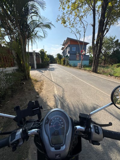 The image depicts a first-person view from the handlebars of a Honda scooter, stationary on a paved shoulder adjacent to an asphalt road in San Phra Net, Thailand. In the immediate foreground, the scooter's dashboard with a speedometer (marked up to 140 km/h) is visible, along with a black smartphone mount affixed to the handlebars on the left, and a chrome-framed rearview mirror on the right.
The road extends straight into the distance, marked by two solid white lines. To the left of the road, a concrete sidewalk or shoulder transitions into a residential area featuring a fence and dense palm trees and other green foliage. To the right of the road, a multi-story building with a light blue lower section, dark blue middle, and orange roofline is visible, displaying air conditioning units and small commercial signs. A large tree with sparse leaves stands between the building and the road, with an overgrown grassy area further to the right. The sky is clear blue with minimal cloud cover, and strong sunlight casts distinct shadows across the road and shoulder, indicating daytime. No individuals or vehicles other than the scooter from which the image is taken are present.