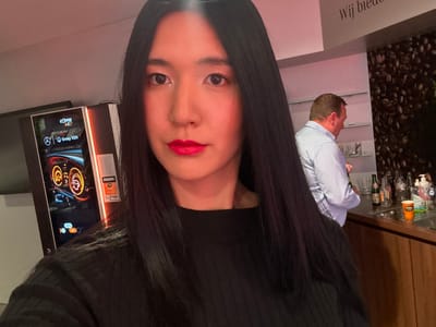 The image depicts an indoor scene within a commercial or corporate environment, specifically inferred to be a Mercedes-Benz Group VDH location in Herentals, Belgium, based on visible branding and Dutch text.

In the foreground, a female with long, straight black hair, fair complexion, and red lipstick is centered, wearing a black ribbed long-sleeved top. She is directly facing the camera.

To her left, in the mid-ground, stands a modern coffee vending machine. Its illuminated screen displays "COFFEE 4 C" and "Mercedes-Benz Group VDH" branding alongside a graphical representation of an automotive dashboard.

In the background, to the right, a male individual with short brown hair, wearing a light blue and white striped shirt, stands partially turned away from the camera. He is positioned behind a wooden bar or counter. On this counter, several bottles (some clear, some green glass), multiple empty glasses, a hand sanitizer bottle, and an orange plastic cup are present. Shelving with additional items is visible behind the counter. A wall section directly behind the bar features a large graphic of coffee beans. Above this graphic, the partial Dutch text "Wij biede" (We offer) is visible.

The overall lighting is artificial and somewhat dim, with a reddish hue present on the left side of the frame. The female is posing for the photograph, while the male appears to be engaged with items on the bar counter. No direct interaction between the two individuals is observable.