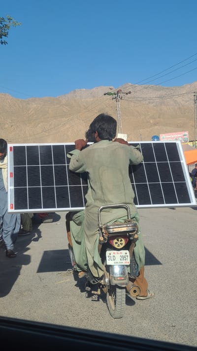 A man is transporting a large solar panel on a motorcycle on a road in Bostan, Pakistan. The solar panel is resting on his back and shoulders. The motorcycle's license plate reads "SLD 2957" with "S.S.D. Sindh 2016" on the plate. The background features a clear blue sky, a mountain range, and utility poles. Another man is standing nearby.