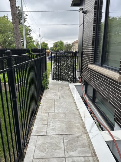 This is a sideways, eye-level shot of a modern building's exterior, featuring a black metal fence and a paved walkway. The building itself is constructed with dark brick and large windows, reflecting the overcast sky and surrounding greenery. A security camera is visible mounted on the building's facade.
The walkway is paved with large, light-colored rectangular tiles that have a subtle marble-like pattern. The black metal fence runs along the edge of the walkway, separating it from a grassy area. Some green plants are growing through the fence, adding a touch of nature to the scene.
In the background, through the fence and trees, a street is visible with a blue vehicle and what appears to be a utility pole with power lines. The sky is cloudy, suggesting it's daytime, possibly in the late morning or afternoon, with diffused light. There is no clear indication of any specific activity or event taking place, as the image appears to be a still observation of the building's exterior and its surroundings. The overall atmosphere is neutral, with no strong emotional cues. No visible text is present in the image. The location context of Mississauga, Canada, doesn't provide any specific visual cues within the frame.