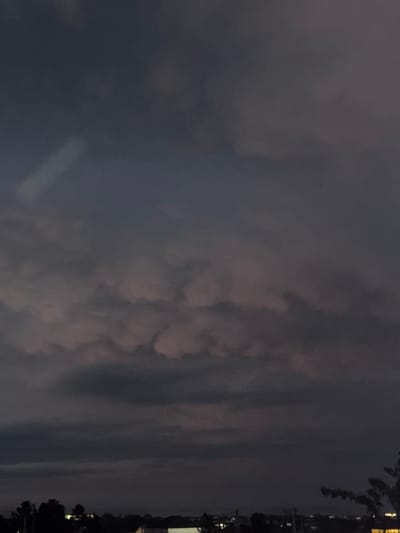 The image shows a night sky with a dark and cloudy atmosphere. A city skyline is barely visible in the distance along the right edge, with some faint lights suggesting urban activity. Silhouettes of trees frame the skyline. There are no visible people or interactions depicted in the image. Based on the prompt, the location is Novxanı, Azerbaijan.