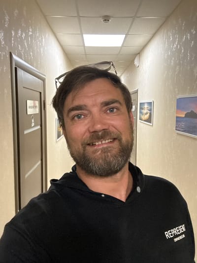 This is a vertical, head-and-shoulders selfie of a man taken in a hallway. The man has brown hair and a beard, and he is smiling at the camera. He is wearing a black t-shirt with white text on the left shoulder that reads "REPRESENT OWNERSCLUB." There are two framed pictures on the wall behind him, and a door with a sign on it. The lighting suggests it is daytime. The scene appears to be indoors, possibly in an office building or institution. The man's expression is happy and relaxed. The text on the door is in Russian and is partially obscured, but appears to say "РЕМОНТ" (REMONTh).