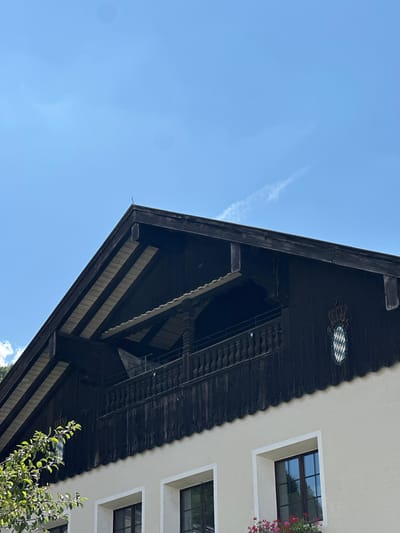 This media file is a low-angle shot looking up at a building with a dark, ornate roof and a light-colored facade. The sky is a clear, bright blue with a wispy cloud. The building appears to be traditional in style, possibly in a mountainous region given the context of Berchtesgaden, Germany.
The subjects are architectural elements of the building. A prominent feature is the dark, wooden overhang of the roof, which has intricate carvings and a scalloped edge. Visible under the eaves are wooden beams and paneling. Above the overhang, on the facade, are at least two windows with dark frames and a white trim. One window has red flowers in a planter box. Above one of the windows, there is a circular decorative emblem with a blue and white diamond pattern. A small tree with green leaves is visible in the lower right corner, partially obscuring the building.
There is no activity or event taking place; the image captures a static scene. The notable details include the time of day, which appears to be daytime due to the bright sky. The weather is clear and sunny. Location cues are the architectural style, the decorative emblem, and the provided location context of Berchtesgaden, Germany. No emotions are conveyed as there are no people present. There is no visible text in the image.
