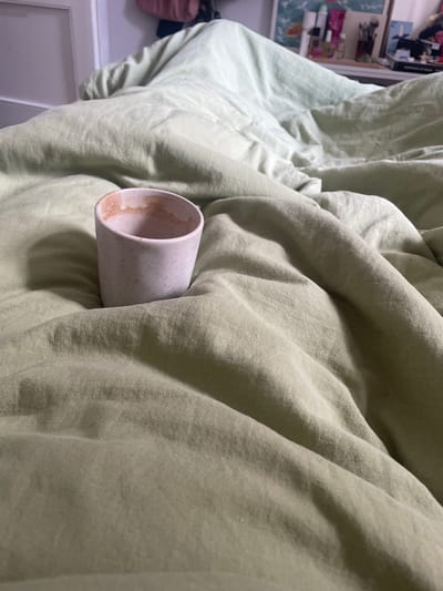 The image displays an indoor scene, specifically a rumpled bed covered with light green linen bedding, located in the city of Piacenza, Italy. A light pink, speckled ceramic cup, tapered at the base and wider at the rim, is centrally positioned on the linen. The interior of the cup contains dried, light brown residue, indicative of a recently consumed beverage. The linen sheets exhibit numerous folds and creases, suggesting recent occupancy or activity. In the blurred background, to the upper left, a white door frame abuts a light-colored wall. To the upper right, various indistinct objects including bottles, what appears to be a brush holder, and other items are visible on a surface, likely a dresser or shelf. A soft, indirect light illuminates the scene, creating subtle shadows within the folds of the fabric and around the base of the cup. No individuals are present.