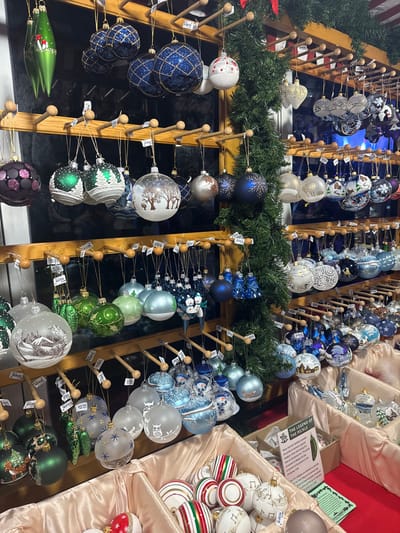 A display of Christmas ornaments features various colors and designs. Blue, white, green, and purple ornaments hang on wooden racks adorned with evergreen garlands. Some ornaments have intricate patterns, while others feature winter scenes. Additional ornaments in white, gold, red, and green fill beige-lined shelves. Small price tags dangle from the ornaments.