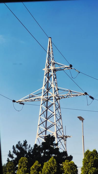 This is a vertical, low-angle outdoor photograph of a large, lattice-style electricity pylon against a clear blue sky. The pylon is made of white metal and has a prominent cross-arm structure from which power lines are suspended. Several black electrical wires are visible, stretching across the frame. Below the pylon, a dense line of green trees, including pine trees and other conifers, forms the lower edge of the image. To the right of the pylon, a modern, gray street lamp stands tall. The scene appears to be in a semi-urban or rural setting. Based on metadata, the image was taken in Ialoveni, Moldova.