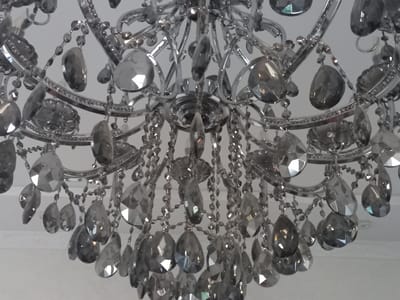 This media file is a close-up, low-angle shot of a crystal chandelier. The chandelier is made of silver-toned metal and features numerous clear and gray faceted crystals of various shapes and sizes, including teardrop and oval forms. These crystals hang from delicate chains, catching the light and reflecting it in bright facets. The chandelier is suspended against a plain, light-colored background, likely a ceiling. The overall impression is one of elegance and opulence. There are no people or specific activities depicted; the focus is solely on the intricate details of the chandelier. The lighting suggests it is daytime, and no particular weather conditions are discernible. The location context of Keles, Uzbekistan, does not provide any specific visual cues within the image itself. There is no visible text in the image.