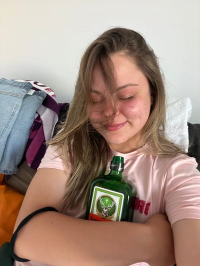A young woman is reclined, possibly lying down, in an indoor setting. She is wearing a light pink t-shirt and has brown hair. She is holding a green glass bottle of Jägermeister. The background shows various clothing items, including a white towel and a blue denim garment. The photo does not provide enough information to determine a specific location in Arroyo de la Miel-Benalmádena Costa, Spain.