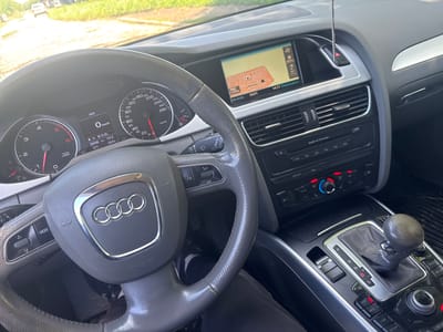 The image captures the interior of an Audi car from the driver's perspective, focusing on the dashboard and center console. Through the windshield, a bright, sunlit road is visible, with a white car in the distance and green foliage lining the sides, suggesting a daytime, outdoor setting.

The prominent Audi steering wheel, featuring the four-ring logo and "AIRBAG" text, is in the foreground. Buttons for "MODE" and volume/track control are visible on the left and right spokes, respectively. Behind the steering wheel is the instrument cluster, displaying two large analog gauges for RPM (left, up to 6 x1000) and speed (right, up to 280 km/h). The digital display between them shows "AUX", "0 km/h", an odometer reading of "206499 km", a trip meter of "741.8", an outside temperature of "+25.0°C", and "D1" indicating the drive gear.

To the right of the steering wheel, an infotainment screen displays a navigation map set to "Brenitsa" with "Nav-Info", an estimated time of "14:17", and "Destination". It also shows "19.5°C" and distances like "400m" and "120m" on the map. Below the screen are climate control vents and the car's sound system controls, including "Audi cd changer" and buttons for "LOAD", "RADIO", and "MEDIA". Further down, the gear shifter is visible, currently in "D" (Drive) mode, with options for "P", "R", "N", "D", and "S". Surrounding the shifter are control buttons for "NAV", "INFO", "CAR", "SETUP", and "RETURN", along with a parking brake button. The overall impression is that of a clean, well-maintained car interior, likely stationary or moving slowly.