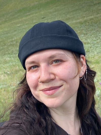 A young person with medium-length wavy brown hair, wearing a black beanie, a brown shirt, a nose ring, and elaborate silver earrings, smiles at the camera outdoors. The background is a blurred grassy hillside with some sparse purple flowers. The person's skin has light freckling, and their eyes are green. The scene is brightly lit, suggesting daylight. The image does not contain any elements to place it in Andreevka, Russia.