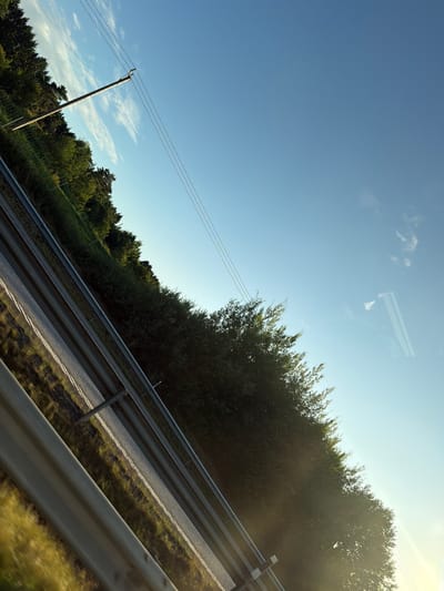 The image shows a view from a car traveling on a road in Kareby, Sweden. The road is bordered by a metal guardrail on the left, with a grassy shoulder and dense trees on the right. A utility pole with three wires is visible on the left side of the road, against a clear blue sky with a few wispy clouds. The sun is low in the sky, casting a warm light and some lens flare. The perspective suggests the photo was taken from inside a moving vehicle, possibly looking out the window.