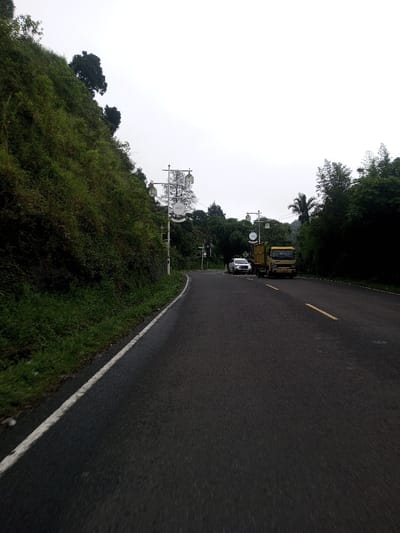 The image displays an asphalt road in Bangtsho, Bhutan, under an overcast sky. The road, appearing dark grey, curves gently to the left in the foreground and straightens in the distance. A solid white line marks the left edge of the road, adjacent to a narrow strip of green grass and weeds. Further down the road, a dashed yellow line indicates a central lane divider. To the left of the road, a steep hillside covered in dense green foliage rises prominently. On the right side of the road, a white four-door sedan is stopped or parked, facing away from the viewer. Directly behind the sedan, a yellow/tan medium-duty truck, also facing away, is stationary. Multiple ornate street light poles with multi-lamp lantern fixtures are positioned along the right side of the road; one pole features a circular decorative element with the inscription "1975". The right side of the road is bordered by dense green trees and vegetation, including a visible palm tree in the mid-ground. No individuals are visible in the scene.