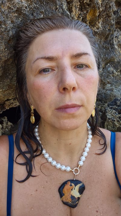 A frontal medium shot shows a woman with wet, dark hair styled back from her face, exposing her forehead and eyes. She is leaning against a rough, grey rock formation, suggesting an outdoor environment, possibly near the sea, given the water droplets on her skin and hair. She is wearing a blue swimsuit, a pearl necklace with a diamond-encrusted clasp, and gold earrings. The necklace features a dark pendant with an illustration of two figures and a swan. The image appears to be taken in the city of Leporano, Italy, perhaps at a coastal location within the commune.