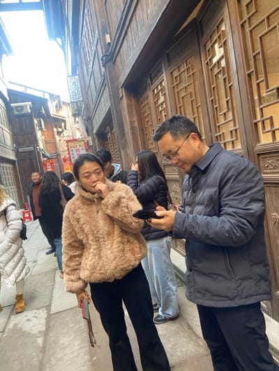 A group of people are walking under a wooden arcade. An Asian man wearing glasses and a dark jacket is at the front. Behind him are two people in dark jackets and blue jeans. A woman with a light brown fur coat is smiling at the camera and holding a phone. More people follow behind. The arcade is ornate with intricate wooden carvings. Buildings are visible in the background. The floor appears to be made of stone.