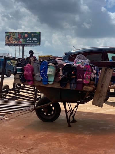 The image shows a wheelbarrow laden with an assortment of flip-flops and sandals, parked on a dusty, reddish-brown surface. The wheelbarrow is positioned on the side of a road, with a blue truck visible in the background to the left. Overhead, a large billboard advertises "ORIJIN NOW IN 33CL." A dark-colored SUV is parked behind the wheelbarrow. A person, wearing a dark head covering and what appears to be a maroon shirt, is partially visible behind the stacked footwear, possibly tending to the merchandise. The sky above is cloudy. This scene is located in Ogbunike, Nigeria.