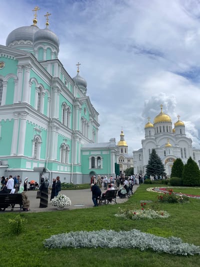 In Diveyevo, Russia, a group of people are gathered on a cloudy day in front of a turquoise and white church with silver domes. To the right, a large white church with golden domes is visible. The people are dressed in modest clothing, many with headscarves, and some are sitting on benches. A pathway is visible between the churches, lined with green grass and flower beds. Several dark green evergreen trees are planted in front of the white church.