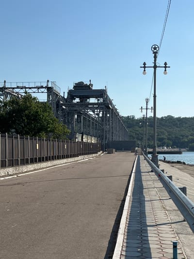 The image depicts an industrial scene with a large metal structure, possibly a conveyor or crane, dominating the left side. This structure is a complex assembly of beams, platforms, and machinery, suggesting a place of industrial activity. To the right of this structure is a paved walkway or road, bordered by a decorative stone pattern. Beyond the walkway, a metal railing runs along the edge of what appears to be a pier or embankment, with the calm blue water of a harbor or sea visible in the background. A lush green hillside covered in trees rises beyond the water, creating a natural contrast to the industrial elements. The sky is a clear, bright blue, indicating a sunny day. The presence of streetlights on tall poles suggests this is a developed area, possibly a port or industrial zone. No people are visible in the frame. The overall impression is one of a functional, outdoor industrial setting on a clear day.