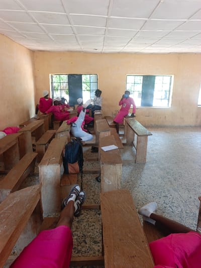 The image depicts an indoor scene within a room located in the City of Mada, Nigeria. The room features light brown, weathered walls with visible chalk markings and a white, grid-patterned ceiling. Two rectangular windows with dark wooden frames are present on the far wall, through which green vegetation is visible. The floor is a light-colored terrazzo, exhibiting signs of wear.

Eight individuals are discernible within the room. Seven are dressed in long-sleeved, bright pink garments, with several also wearing white head coverings. One individual wears a white long-sleeved top and light-colored lower garments.

In the immediate foreground, the legs and lower torsos of two individuals are visible. On the left, legs in a pink garment are shod in dark sandals with white socks. On the right, legs in a pink garment wear white socks, with feet resting on a wooden bench.

In the mid-ground, multiple individuals are seated on simple, unvarnished wooden desks and benches, which are arranged in rows and show signs of use. A person in white attire is seated on a bench, with one hand raised, holding a white object. To their right, another individual in a pink garment sits on a desk, holding a dark object to their mouth, and displays a white identification badge on their chest. Further back, near the windows, an individual with dark hair, wearing a white shirt, is seated on a desk.

A dark-colored backpack with orange accents is placed on the floor between rows of benches in the mid-foreground. White papers are visible on some of the desks.