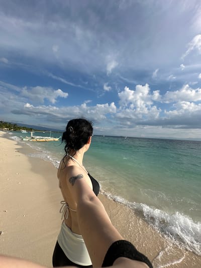 A person with dark hair, facing away from the camera, stands on a sandy beach. They are wearing a black bikini top with white straps and a white sarong tied around their waist over black bottoms. A butterfly tattoo is visible on their left upper arm. The person is holding the camera in their right hand, as if taking a selfie.
The background features the ocean with clear, turquoise water and gentle waves lapping the shore. Several outrigger boats are visible in the shallow water, and a few are anchored further out. In the distance, a coastline with palm trees and vegetation is discernible, suggesting a tropical location. The sky above is a vibrant blue with scattered white, fluffy clouds.
The scene appears to be taken in Saavedra, Philippines, a location known for its beautiful beaches and clear waters, making it a popular spot for water activities and relaxation. The lighting suggests it is daytime, possibly late afternoon given the angle of the sun.