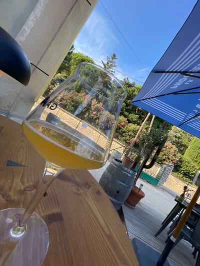 An outdoor table setting with a glass of beer on a wooden table is shown in the foreground, with a bright blue umbrella and lush greenery in the background. The glass, which is tilted, contains a light-colored beer with a frothy head. The table appears to be made of aged wood with visible grain. Behind the table, a wooden barrel and potted plants are visible, suggesting a rustic or garden setting. A blue umbrella with white stripes provides shade, and beyond that, a fence, trees, and a stone wall can be seen. The sky is clear and blue, indicating a sunny day. The scene is set in Cucuron, France. No people are present in the image.