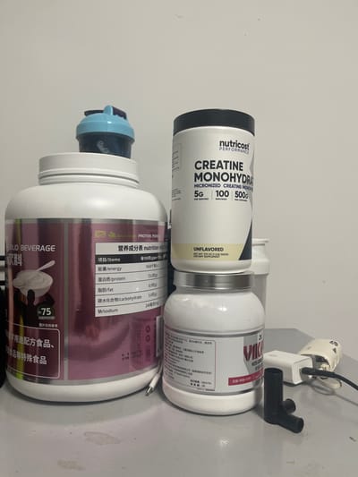 The image displays an indoor scene, likely on a light-colored table or surface against a plain wall. The photo is rotated 90 degrees clockwise, showing objects on their sides.

The primary subjects are three containers of nutritional supplements. In the foreground, a large white tub with a purple and white label is visible. Its label reads "PROTEIN POWDER" and "SOLID BEVERAGE 饮料" (beverage) in English and Chinese. A "营养成分表 nutrition info" (nutrition information) panel lists "能量/energy", "蛋白质/protein", "脂肪/fat", "碳水化合物/carbohydrate", and "钠/sodium" with corresponding values per 100g. The label also shows "~75 SERVINGS," "图片仅供参考" (Image for reference only), and "用途配方食品、限食品等特殊食品" (Special foods for formula food and restricted food use). A blue shaker bottle lid rests on top of this container.

Behind and to the right, a white container with a black lid from "nutricost PERFORMANCE" is prominently featured. Its label specifies "CREATINE MONOHYDRATE MICRONIZED CREATINE MONOHYDRATE," indicating "5G PER SERVING," "100 SERVINGS," and a total of "500G." It is also marked "UNFLAVORED DIETARY SUPPLEMENT 1000G."

Adjacent to the Nutricost container, another white container with a silver lid is partially visible, displaying text like "VIK," "VIES-CHO," "GRONTSA," and some Chinese characters.

In the upper right background, an electrical adapter is plugged into a wall socket, with a black cable and a distinct black Y-shaped plastic piece nearby. The presence of Chinese text on multiple labels is consistent with the Shenhe District, China context. The lighting suggests a typical indoor setting.