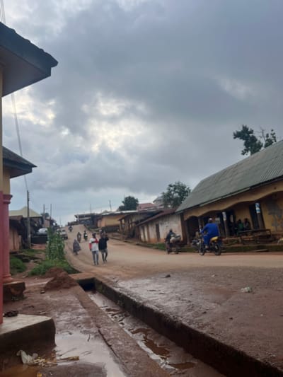 The image depicts a street scene in Gembu, Nigeria, under a cloudy sky suggesting either early morning or late afternoon. The street is unpaved and appears to be wet, with puddles indicating recent rain. Buildings line the street, with some showing signs of wear and tear.
Several people are visible, some walking and others on motorcycles. One person in a blue garment is on a motorcycle, while others are seen in groups or individually on the street. There are multiple motorcycles present, suggesting they are a common mode of transportation in this area. Greenery is visible in patches along the street, indicating some vegetation.
The overall atmosphere is one of everyday activity, with people going about their business despite the overcast weather. The lighting is subdued due to the clouds, and there is no clear indication of any specific event. The wet ground and puddles are a prominent detail, hinting at the tropical climate of the region. No visible text is discernible in the image. The composition of the photograph, with its tilted perspective, adds a dynamic feel to the scene.