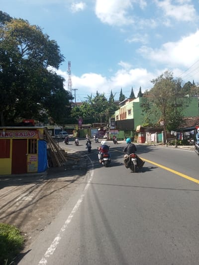 This image captures a daytime street scene in Ban Phonhxavanh, Laos, under a clear blue sky with scattered white clouds. The paved asphalt road features a solid yellow center line and a white line marking the left shoulder. At least ten motorcycles and scooters, most with riders wearing helmets, travel away from the viewer. The foremost motorcycle carries a single rider in a blue helmet, with a brown object secured over the rear seat.

To the left of the road, a dirt shoulder borders a yellow and red structure, identified by signage as a "MIE GORENG BALAP" food stall. Adjacent to this stall, a pile of wooden planks and a blue tarp are visible. A large, dense green tree obscures part of the background, behind which a red and white cellular communication tower stands tall, accompanied by utility poles and overhead wires.

On the right side of the road, a series of buildings line the street. A multi-story building, predominantly light green with brown trim, is visible. Further down, another building displays clothes hanging outside. Signage, including "ASIA OPTIK," is present on some structures. Conical evergreen trees are visible on the upper right, above the buildings. Sunlight casts distinct shadows across the road and roadside elements.