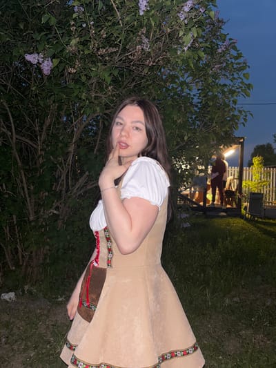 A woman with long, dark hair and light skin poses outdoors in Пальники, Russia, wearing a traditional dirndl dress with a white blouse and tan overall. She touches her finger to her lips, gazing towards the camera. Lilac bushes are on the left, with a group of people under a lit structure in the background, and a grassy area extends towards the right. It appears to be twilight or night.