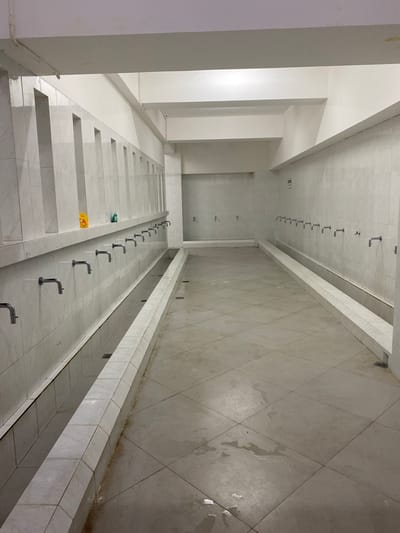 This is a wide-angle, eye-level, indoor photograph of a communal washing area, likely in Aluekambuk, Indonesia. The room is brightly lit with overhead lighting. The walls and floor are tiled with large, light-colored square tiles. Along the left wall, there is a row of chrome faucets mounted on the tiled wall, above a long, trough-like basin. These basins are also lined with tile and appear to have drains. Further down the room, along the right wall, there is another, identical setup of faucets and basins. The floor is mostly dry but shows several wet patches and puddles, indicating recent use. The overall impression is of a functional, clean, and well-maintained public facility. There are no people visible in the image.